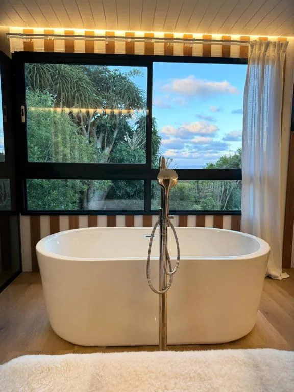 Freestanding oval bathtub with handheld shower, garden and sky views through large windows