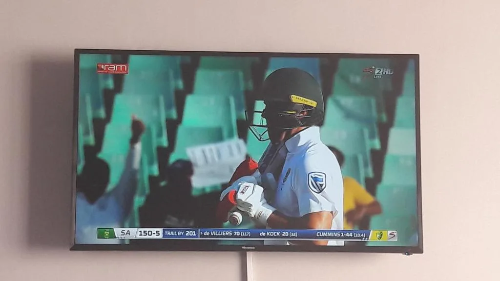Flat-screen television mounted on wall displaying cricket match broadcast