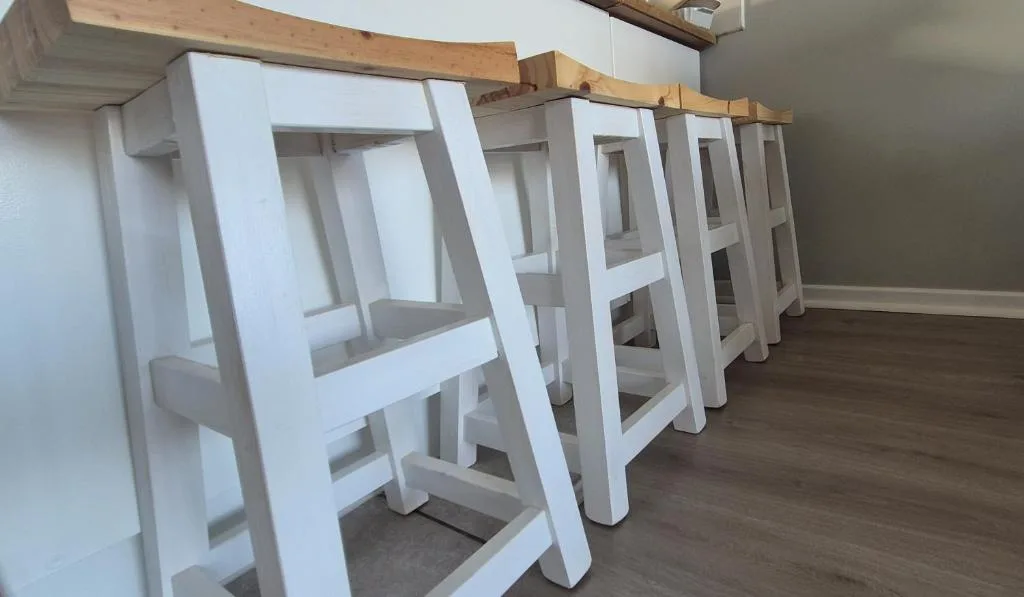Row of white wooden bar stools with natural wood tops lined along counter