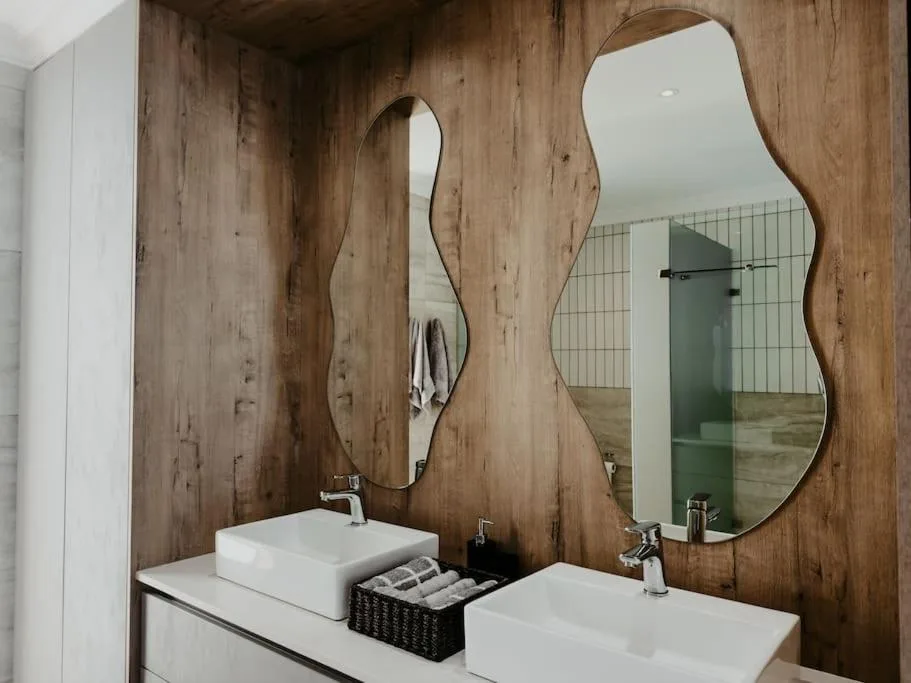 Modern bathroom with dual white sinks, wooden wall paneling, curved mirrors