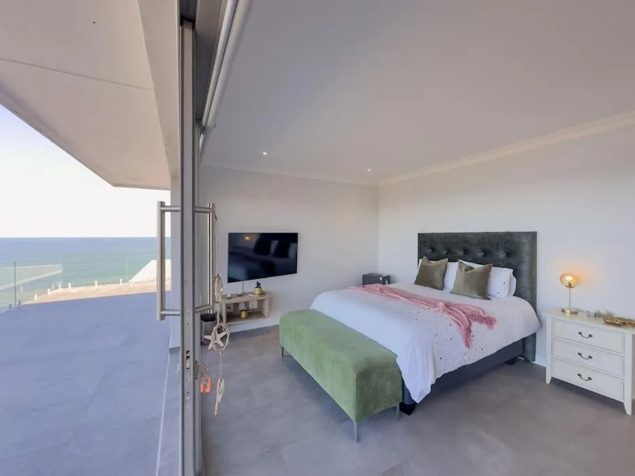 Luxury bedroom with ocean view, bed with dark headboard, green bench, and open deck doors