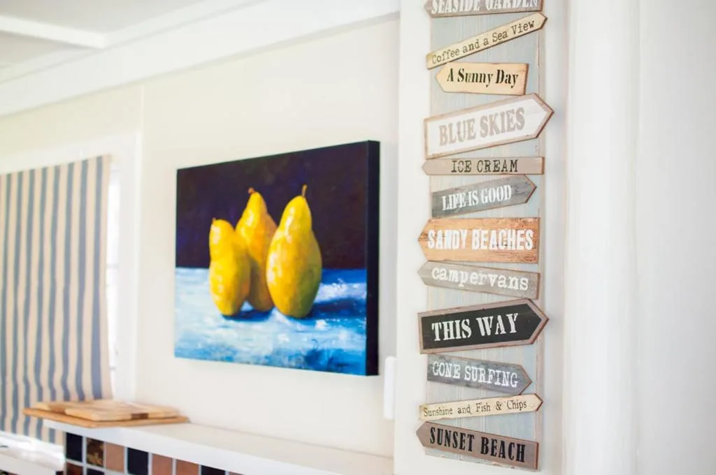 Coastal-themed wooden sign stack and yellow pear artwork on white wall