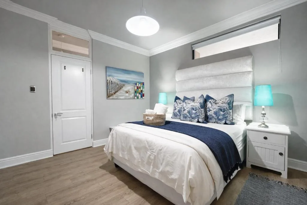 Bright coastal-themed bedroom with queen bed, turquoise lamps, and seaside artwork