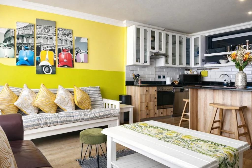 Bright yellow lounge with striped sofa, colorful Vespa wall art, and open kitchen