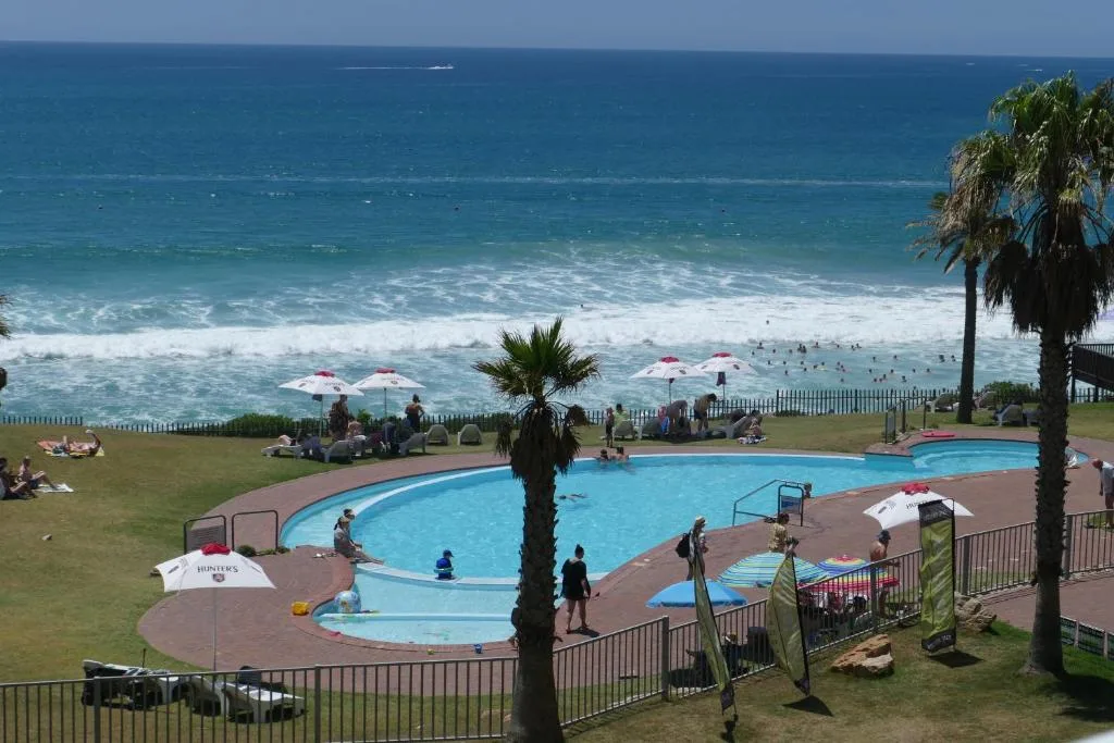 Curved swimming pool with beach and ocean view beyond, palm trees, umbrellas