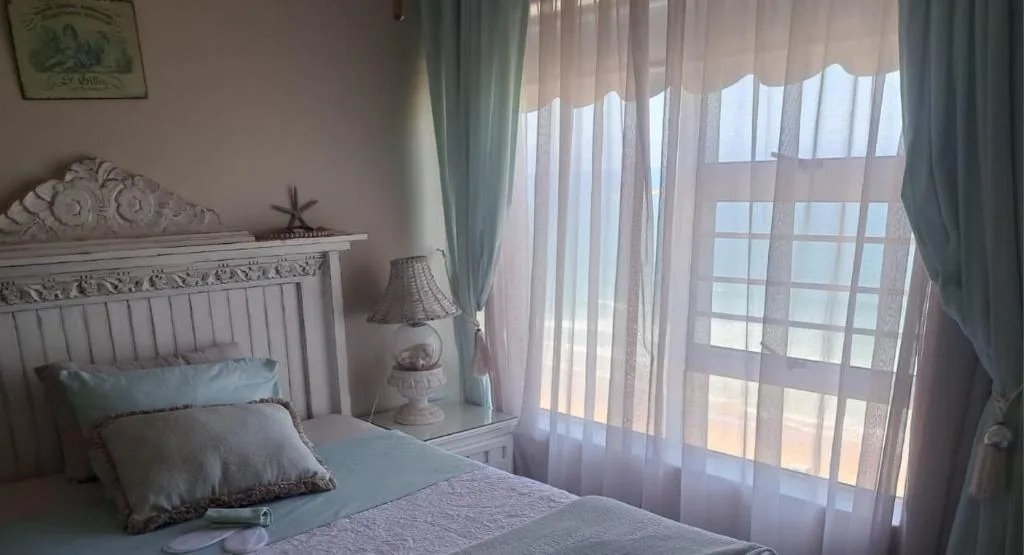 Bright bedroom with white bed frame, blue pillows, and sheer curtains overlooking ocean view