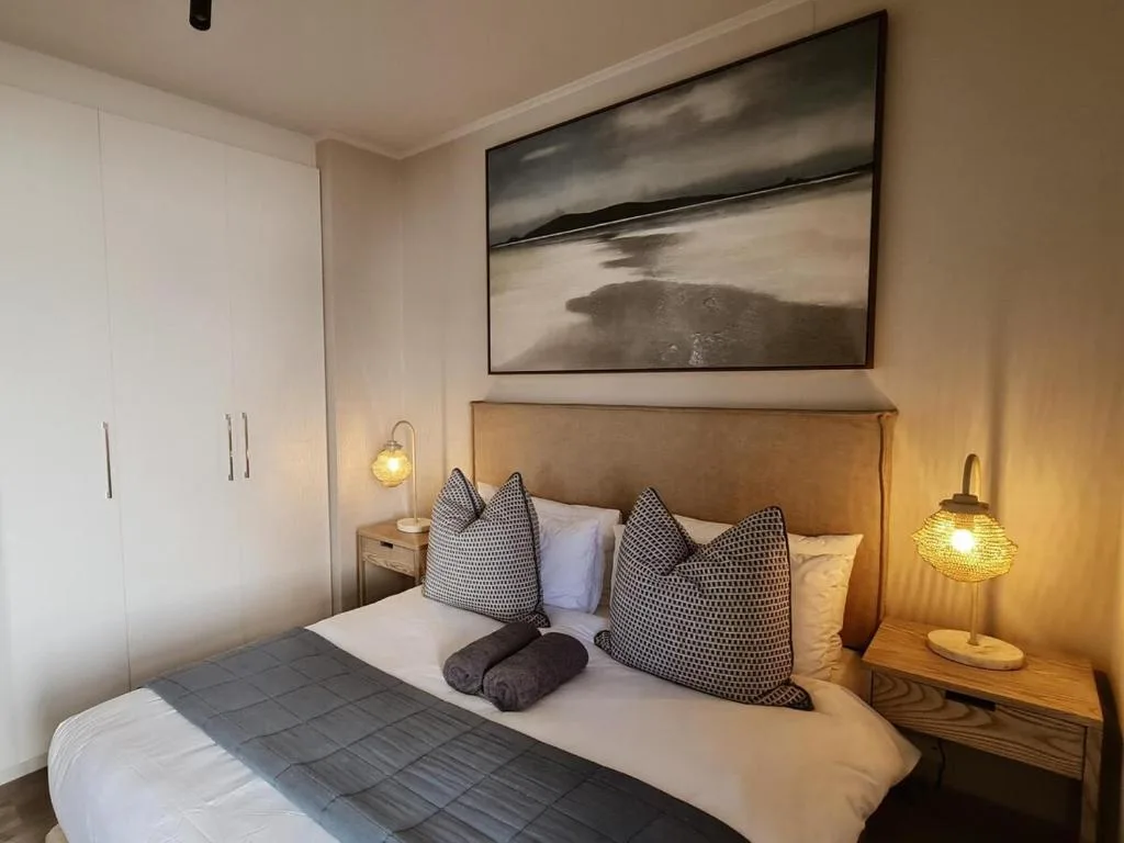 Modern bedroom with tan headboard, patterned pillows, and warm bedside lighting