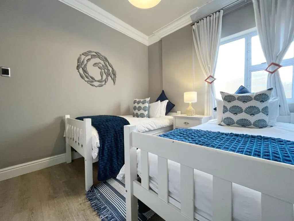 Bright bedroom with two white beds, blue accents, coastal artwork, and large windows