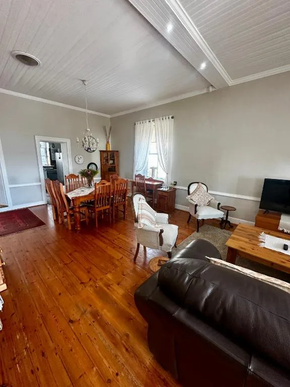 Open-plan lounge with black leather sofa, dining table, wooden floors, and bright windows