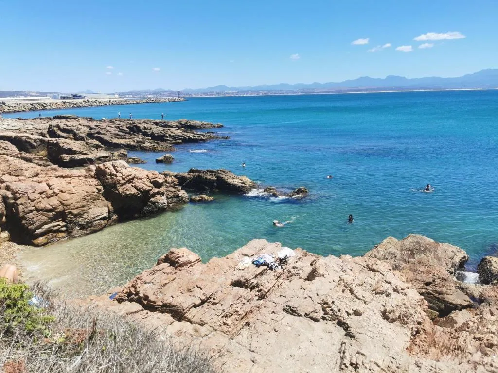 Turquoise ocean cove with rocky coastline and distant mountains under clear sky