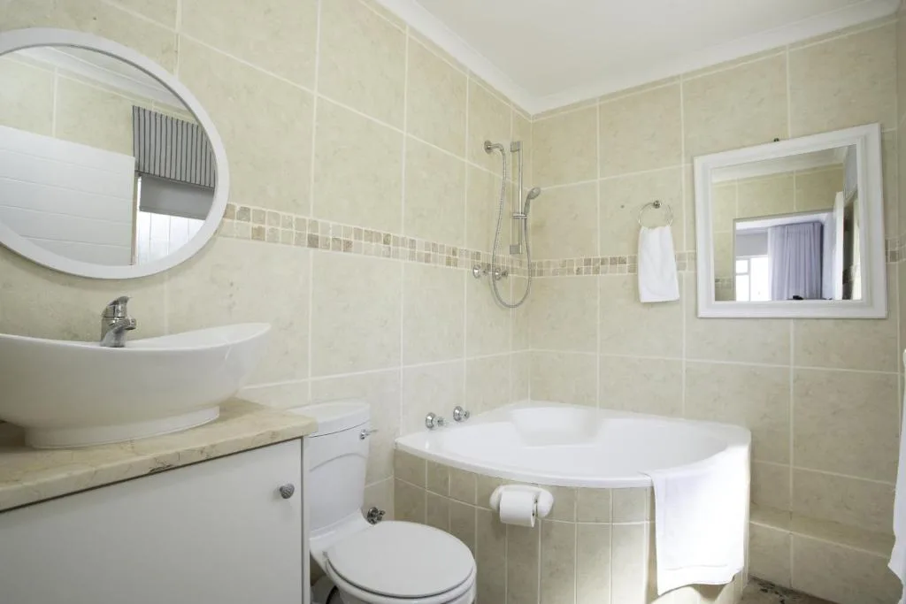 Cream tiled bathroom with round mirror, basin, toilet, and bathtub
