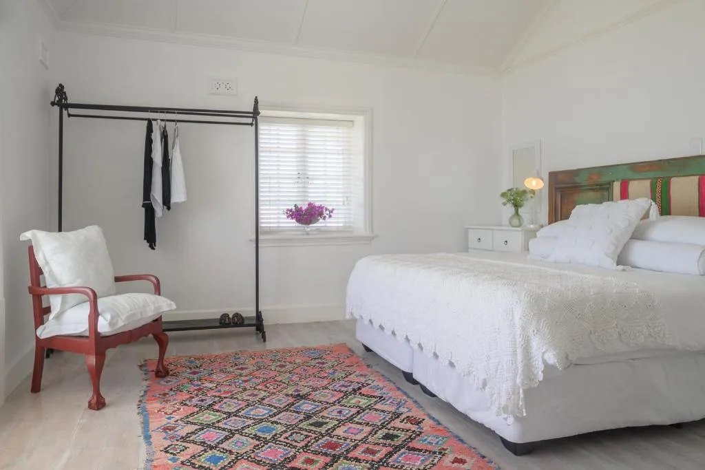 Bright bedroom with white linens, colorful rug, red chair, and bedside flowers
