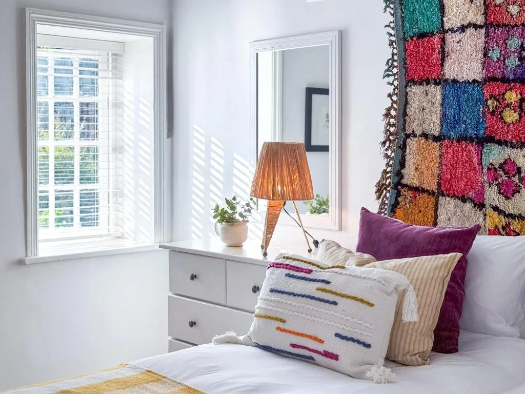 Bright bedroom with colorful pillows, shuttered windows, and textile display