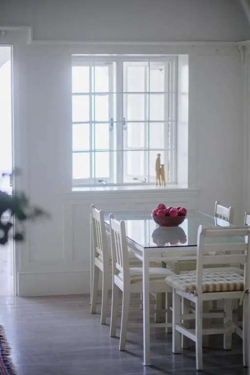 Bright dining room with white chairs and table, large windows overlooking garden