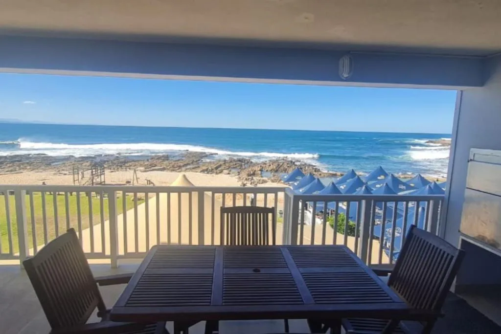 Beachfront deck with dining table overlooking ocean, rocks, and sandy beach.