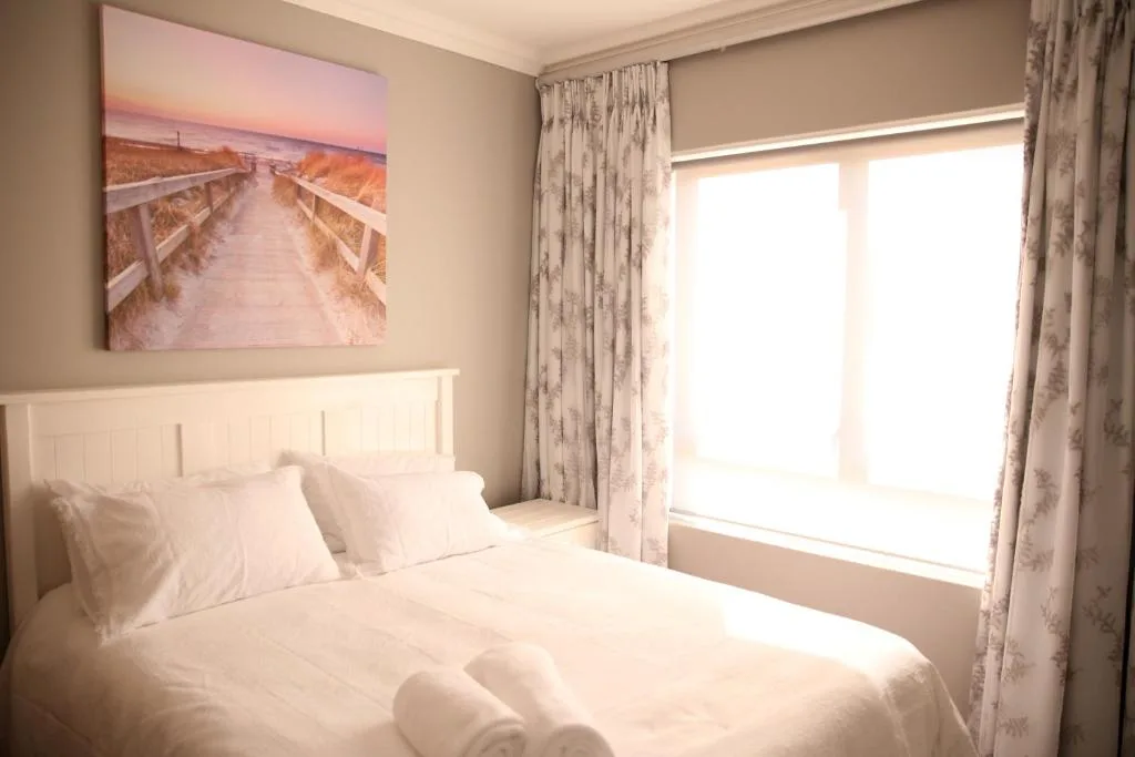 Bright bedroom with white bedding, coastal artwork, and large windows