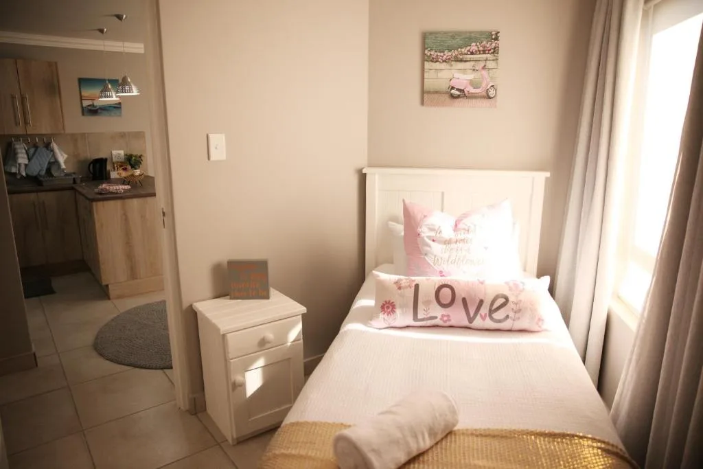 Bright bedroom with single bed, pink cushions, white headboard, kitchenette visible