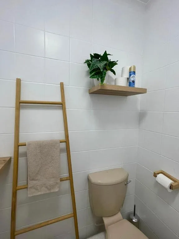 Modern white tile bathroom with toilet, wooden towel ladder, and decorative shelf