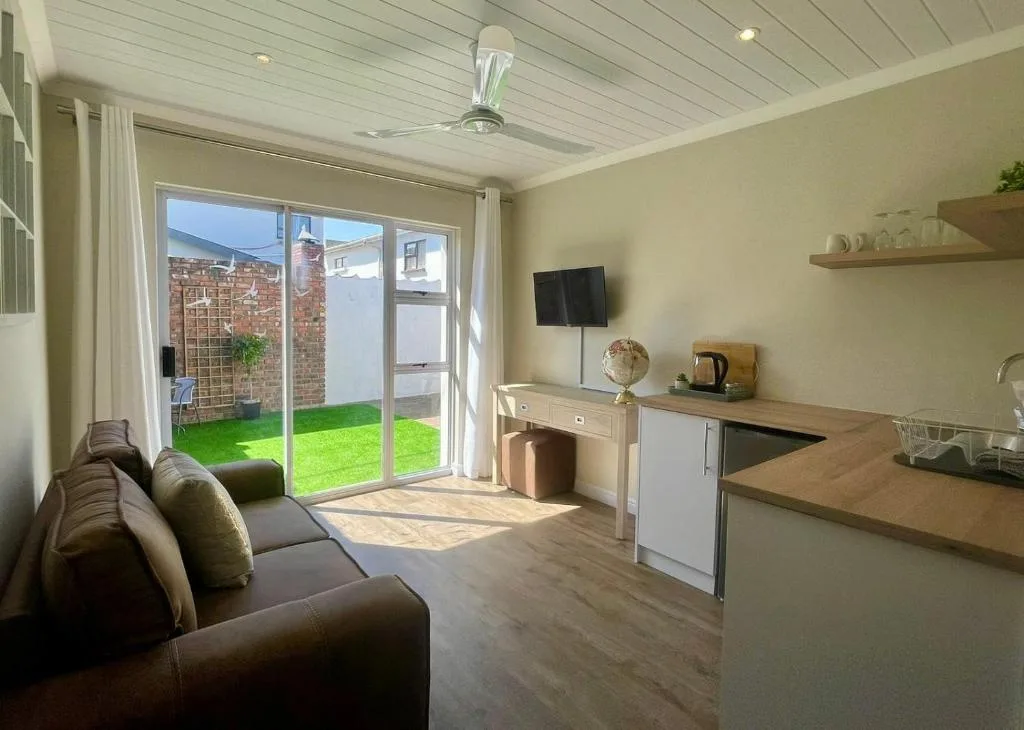 Bright lounge with brown sofa, TV, kitchenette, and garden views
