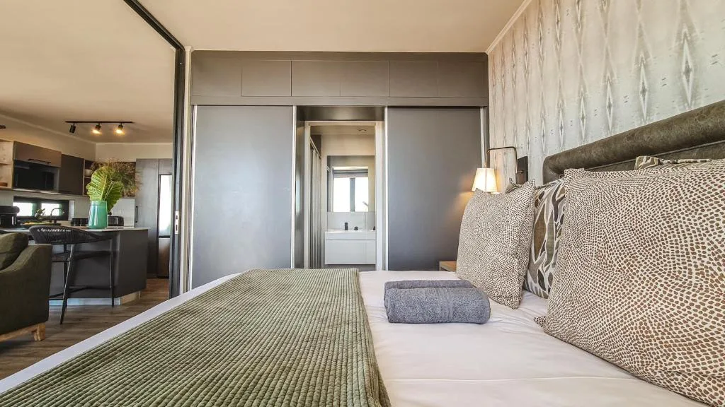Modern bedroom with queen bed, ensuite bathroom visible, open-plan kitchen beyond