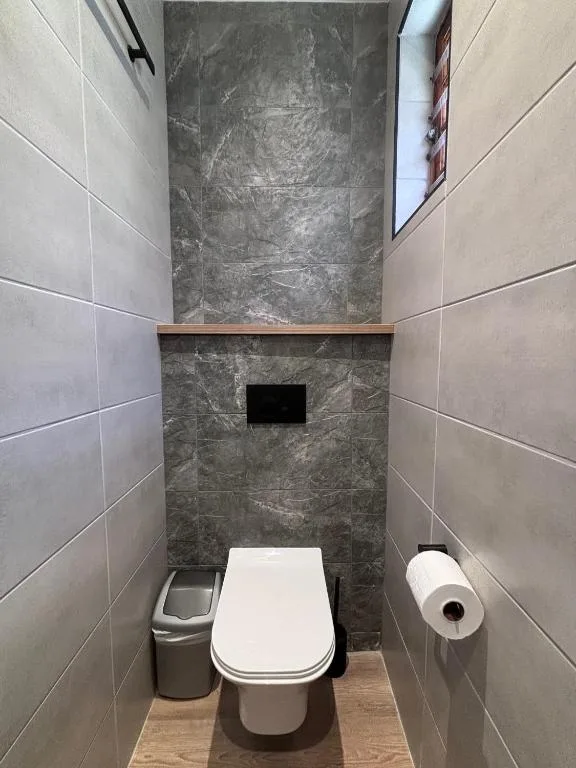 Modern bathroom with gray tile walls, white toilet, and small trash bin