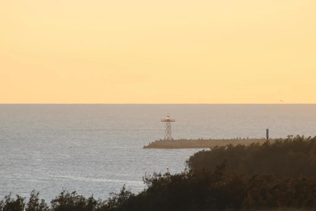 Sunset over ocean with lighthouse and breakwater visible on horizon