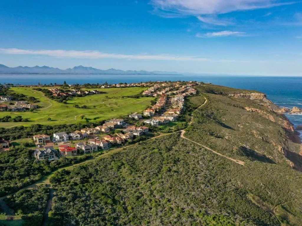 Aerial coastal landscape showing lush green hills, residential properties, ocean, and distant mountains