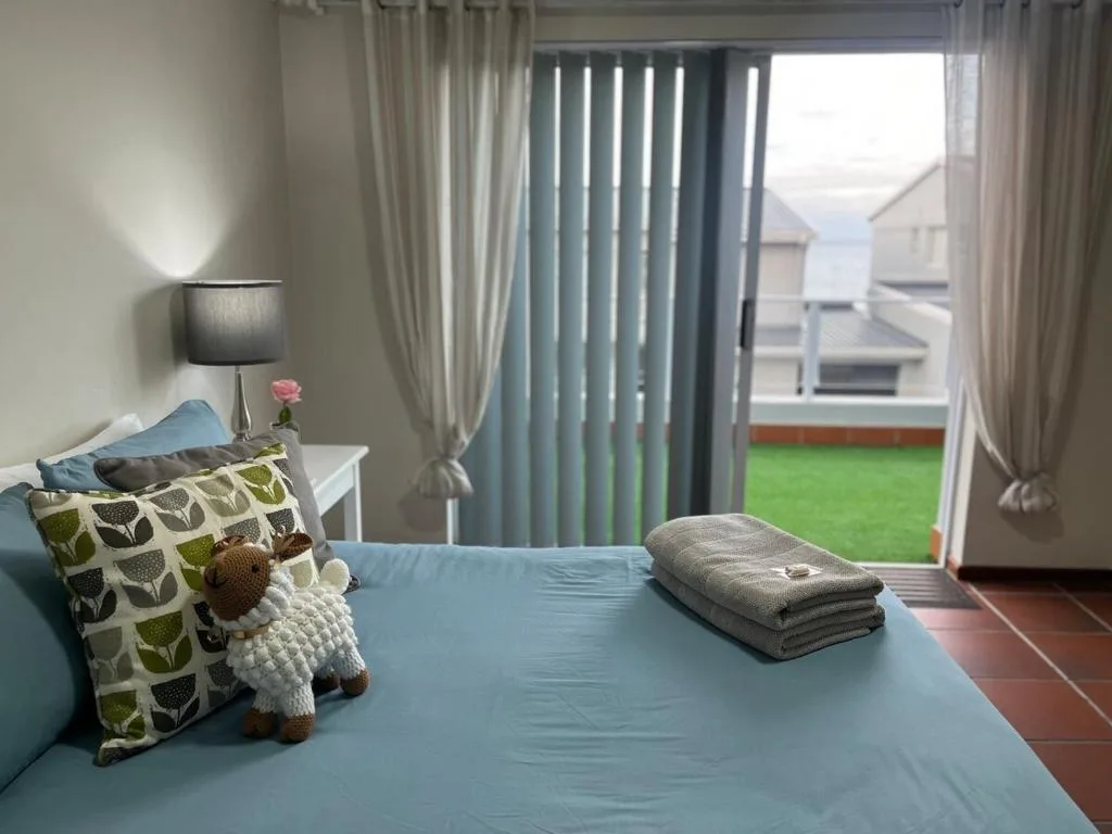 Bright bedroom with blue bedding, decorative pillows, and garden views through vertical blinds