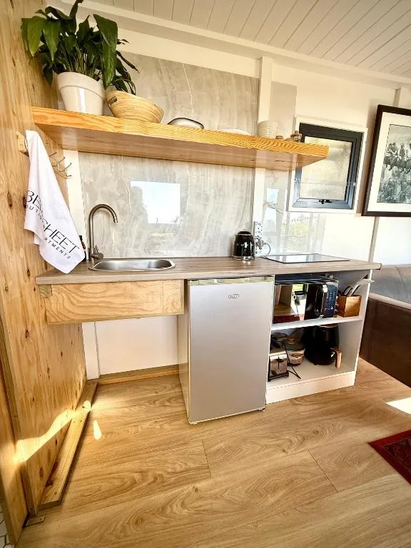 Compact kitchen with sink, mini fridge, wooden shelving, and window view