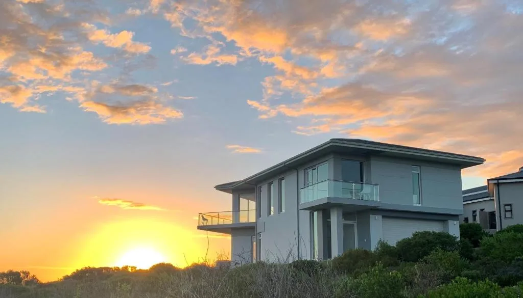 Modern two-story white villa at sunset with coastal vegetation and dramatic sky