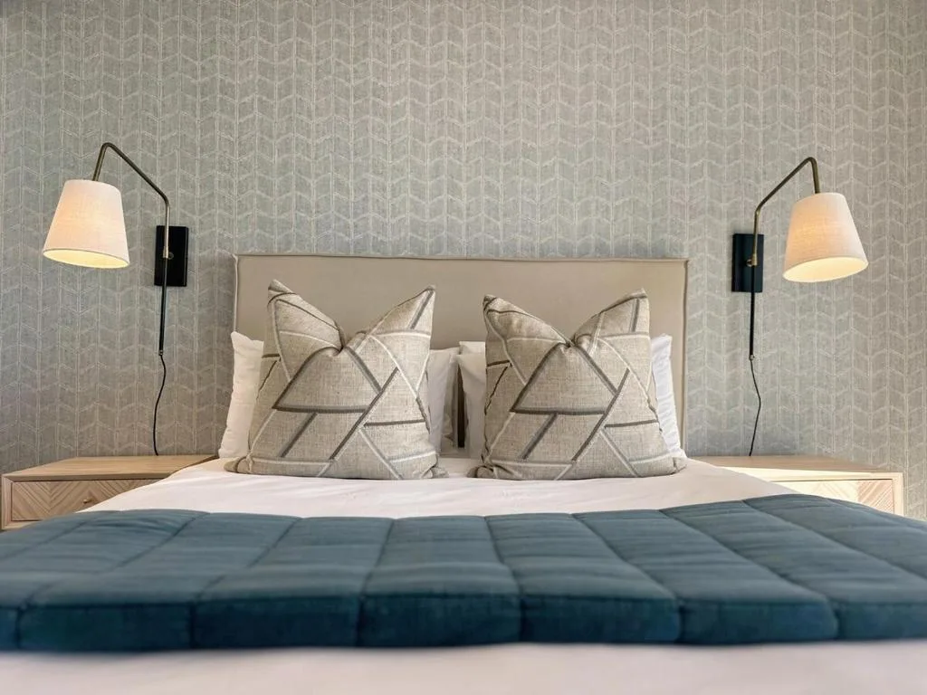 Modern bedroom with upholstered headboard, geometric patterned pillows, dark bedding, and wall-mounted reading lamps