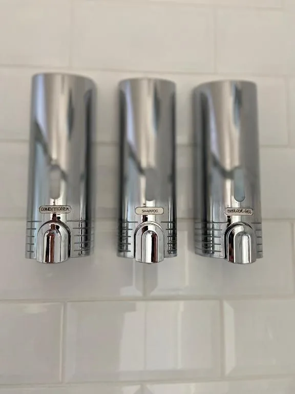 Three chrome soap and shampoo dispensers mounted on white tiled wall