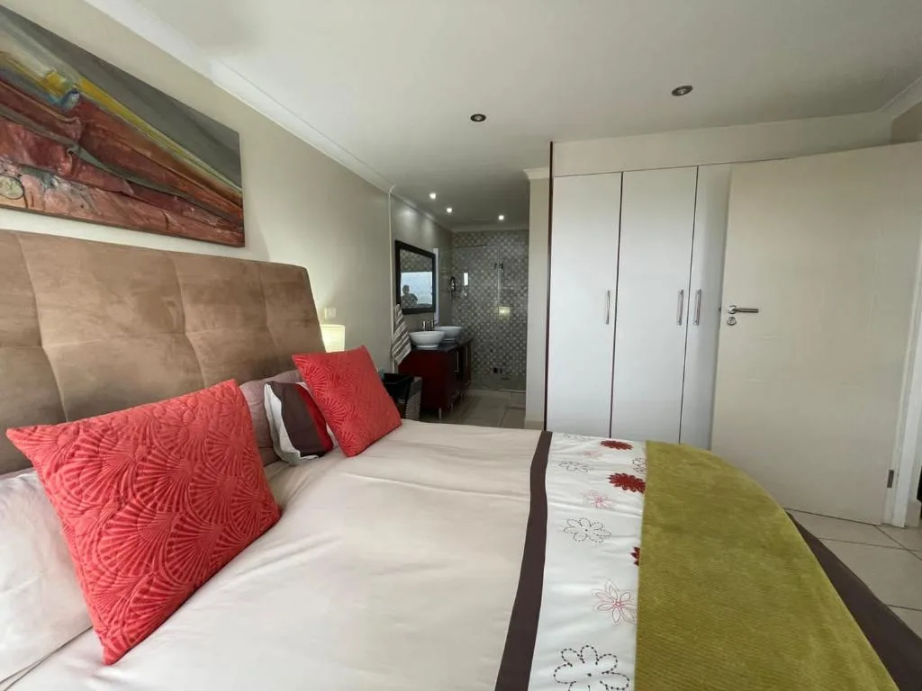 Modern bedroom with taupe headboard, red patterned pillows, ensuite bathroom visible