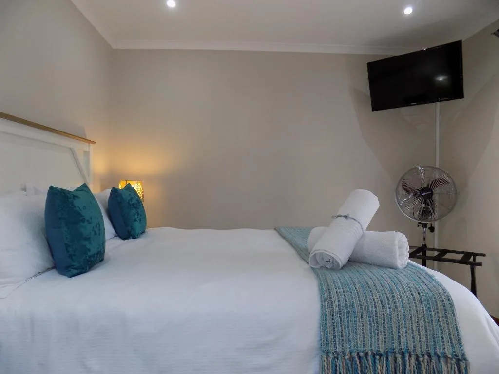 Modern bedroom with white linens, teal cushions, wall-mounted TV, and fan