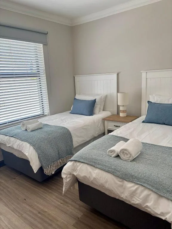 Twin bedroom with white headboards, blue cushions, rolled towels, and shuttered window
