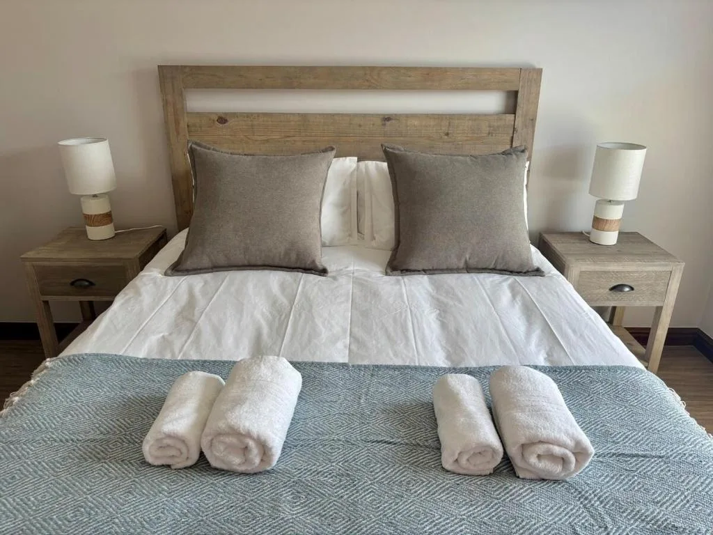 Master bedroom with wooden headboard, grey pillows, white linens, and rolled towels
